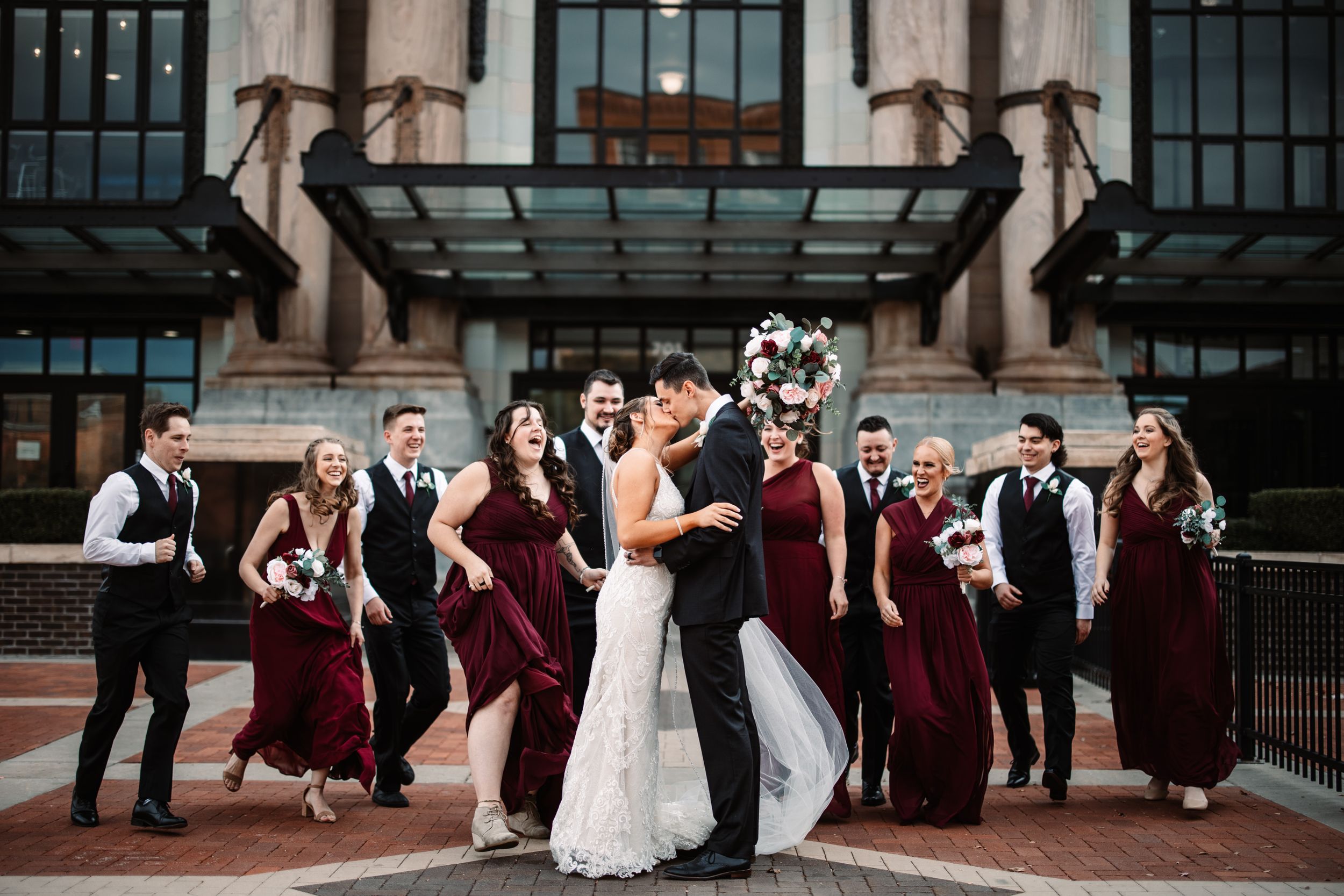 Wedding party in burgundy dresses and black suits pose together on stone steps under an ornate archway.