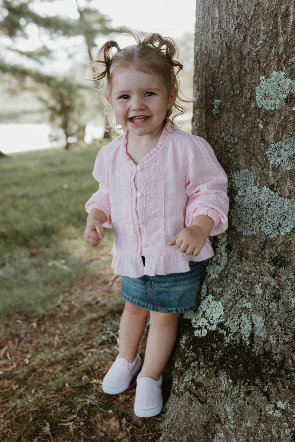 Child in pink shirt and denim shorts sitting by large tree trunk in natural setting.