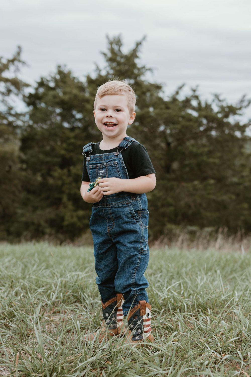 Small child wearing denim overalls walks through tall grass in outdoor setting.