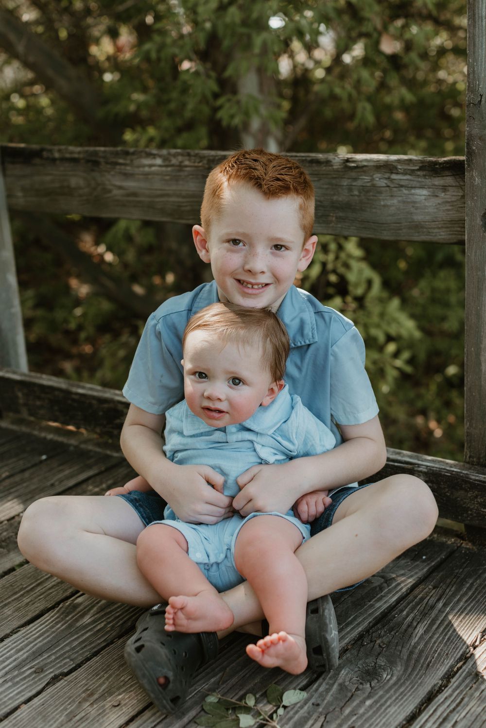 Two children sitting together on weathered wooden deck in outdoor setting.