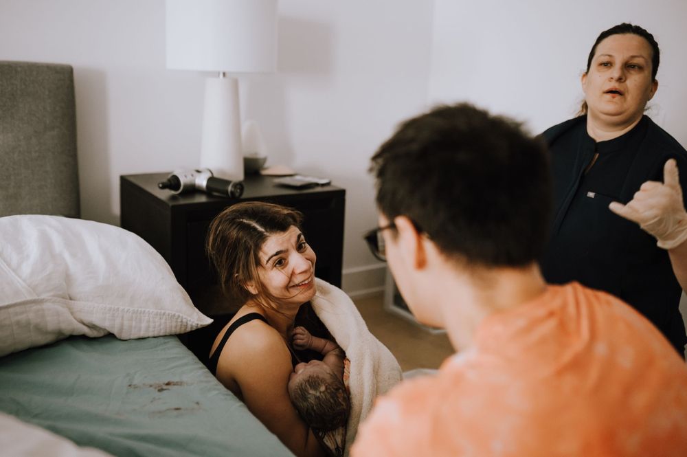 Three people interact in a casual bedroom setting with a white lamp and gray upholstered furniture.