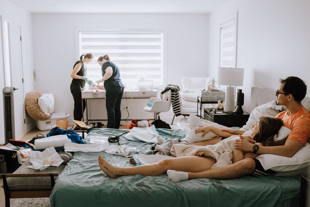 People lounging in a messy bedroom with clothes strewn about while others stand by a window in the background.