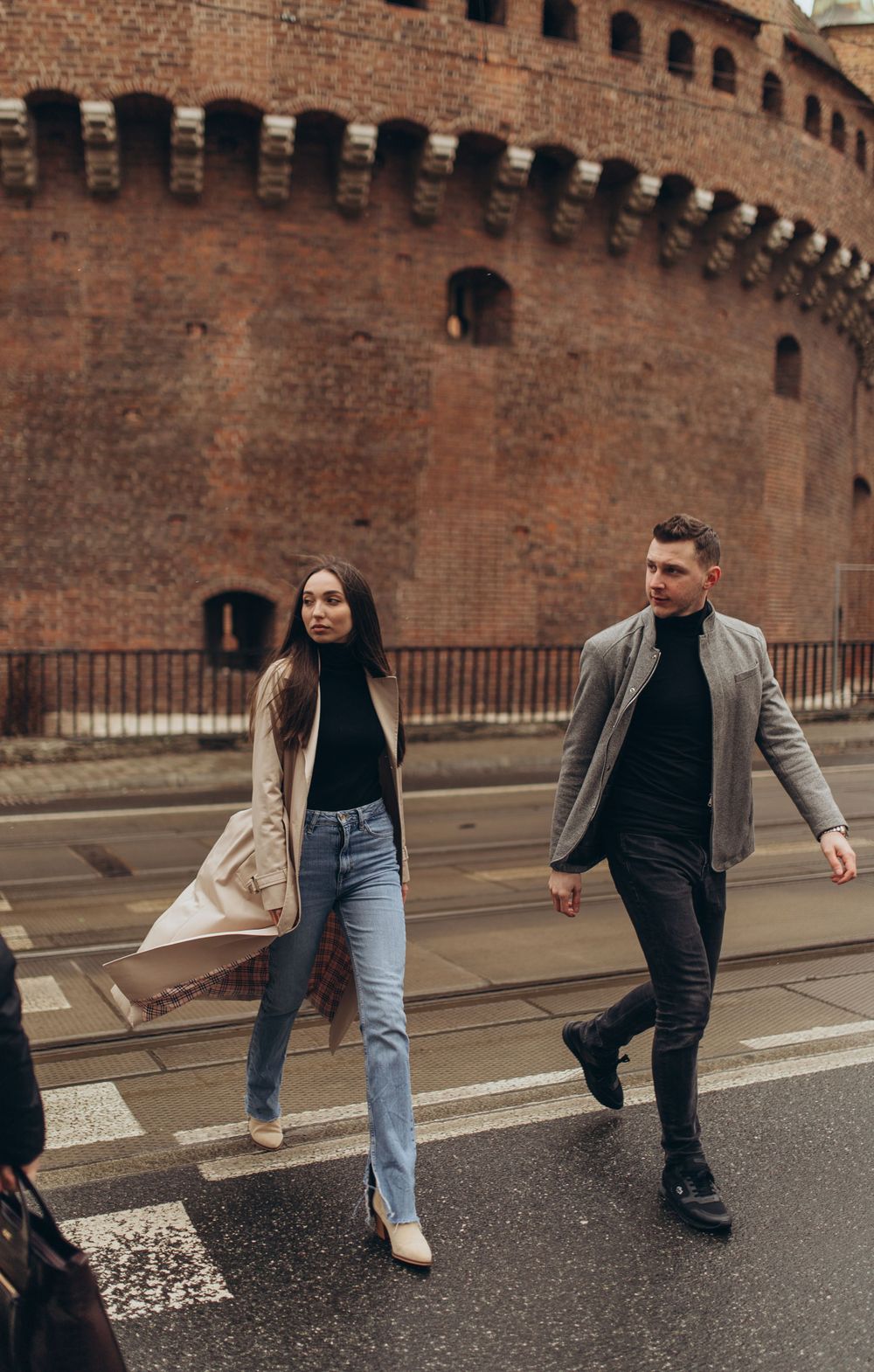 Couple in stylish outfits walking past historic brick fortress wall.