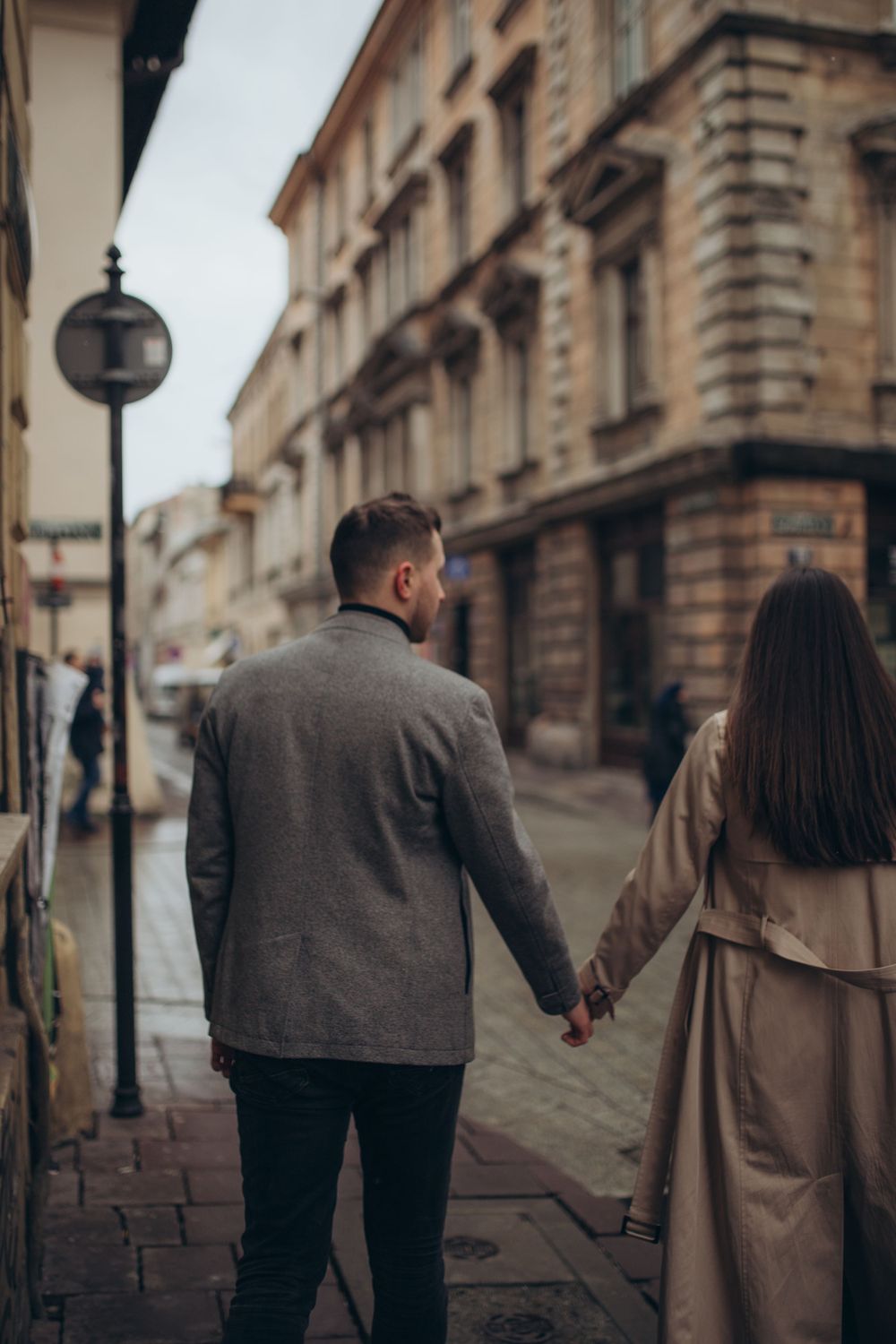 Couple in coats walking hand in hand down a cobblestone street with classic buildings.