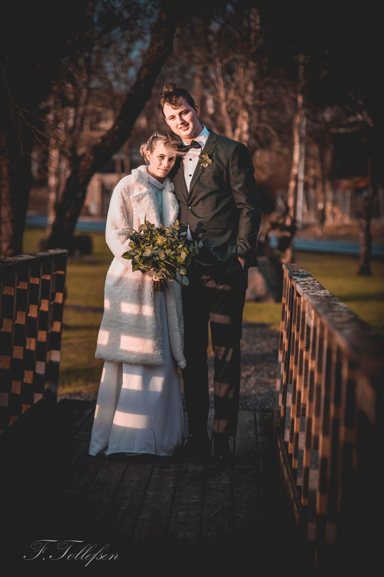 Bride and groom posing together on a bridge in warm sunlight with a green foliage bouquet.