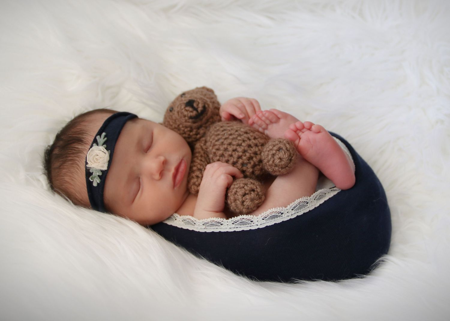 Sleeping newborn swaddled in navy fabric holding a knitted brown teddy bear on white fur backdrop.