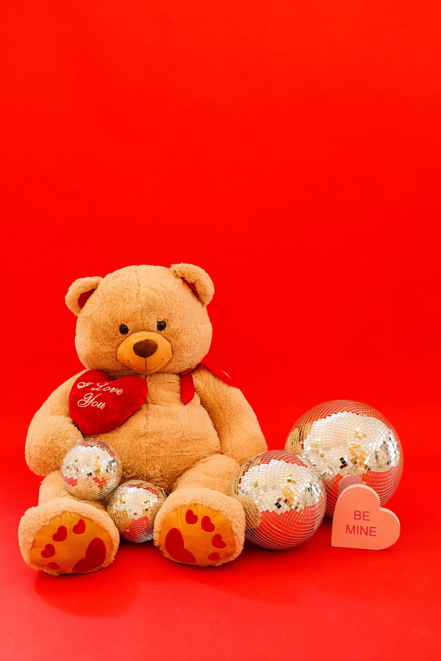 Plush teddy bear holding heart with silver ornaments against bright red background for Valentine's Day.