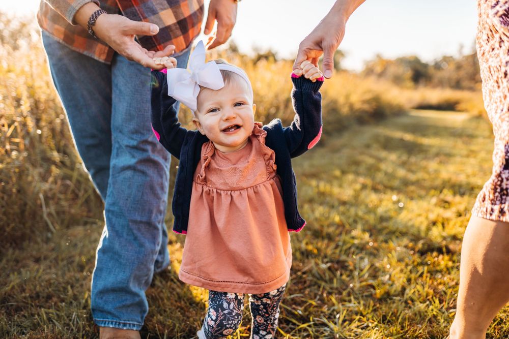 Baby in a peach dress walking with adult support in a sunlit field.