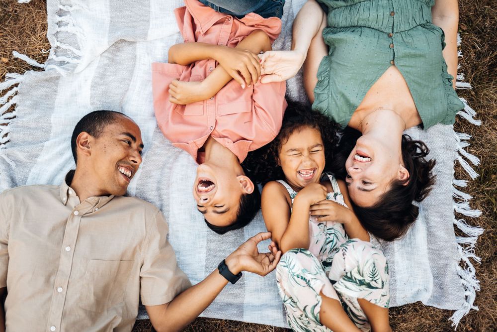 A family lies on a white blanket outdoors laughing and having fun together.