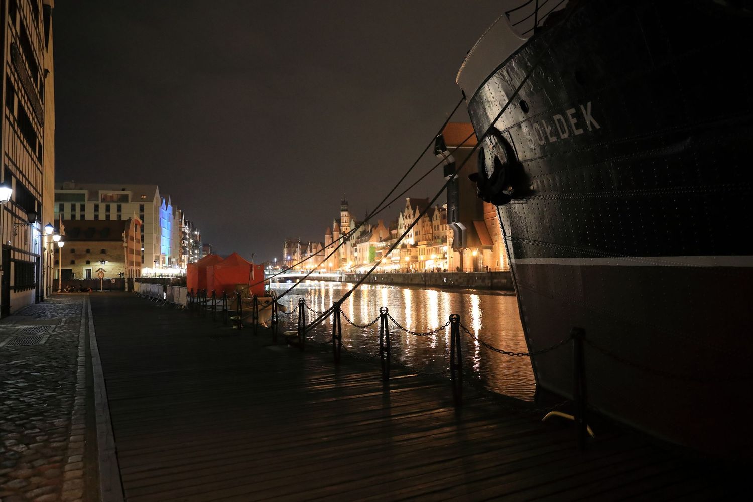 Ships docked along a dimly lit waterfront promenade at night with historic buildings in the background.