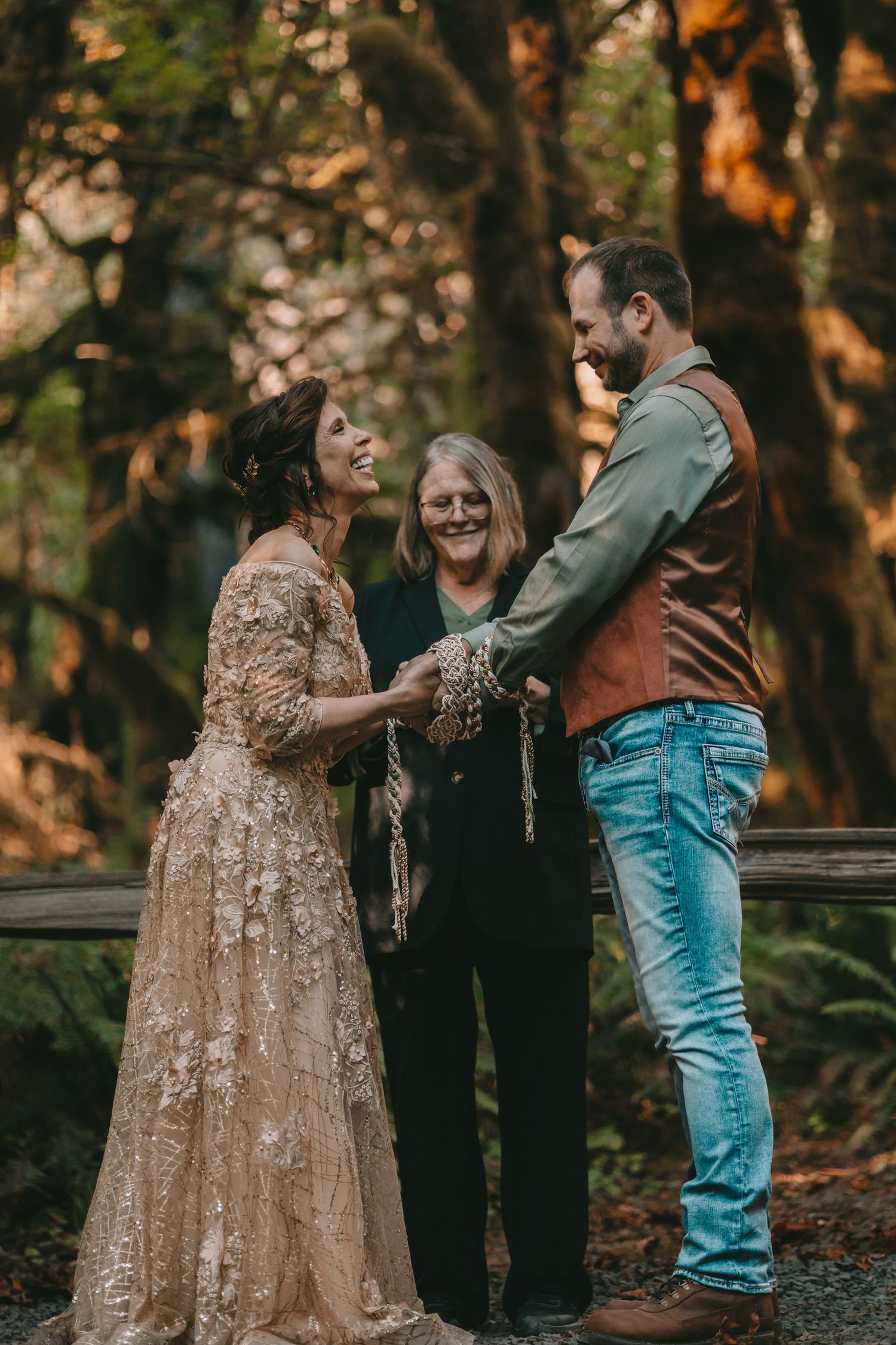 Couple stands together in Hoh Rainforest Olympic national park in wedding ceremony holding hands with rope around officiant behind them