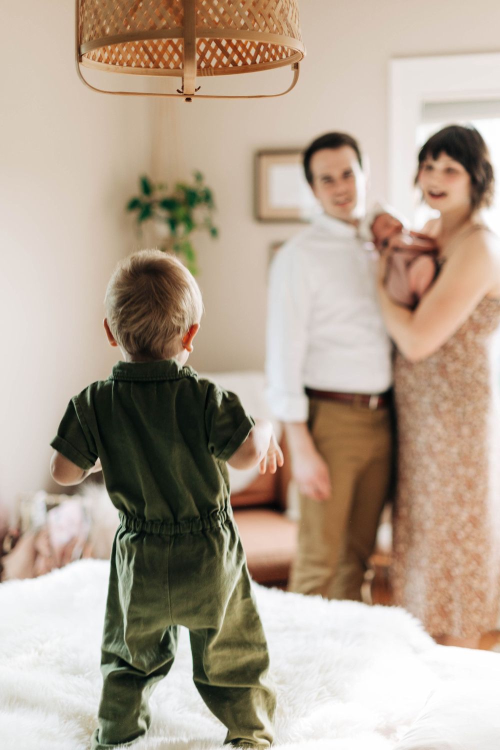 A toddler in green overalls stands on a bed while others interact in the background.