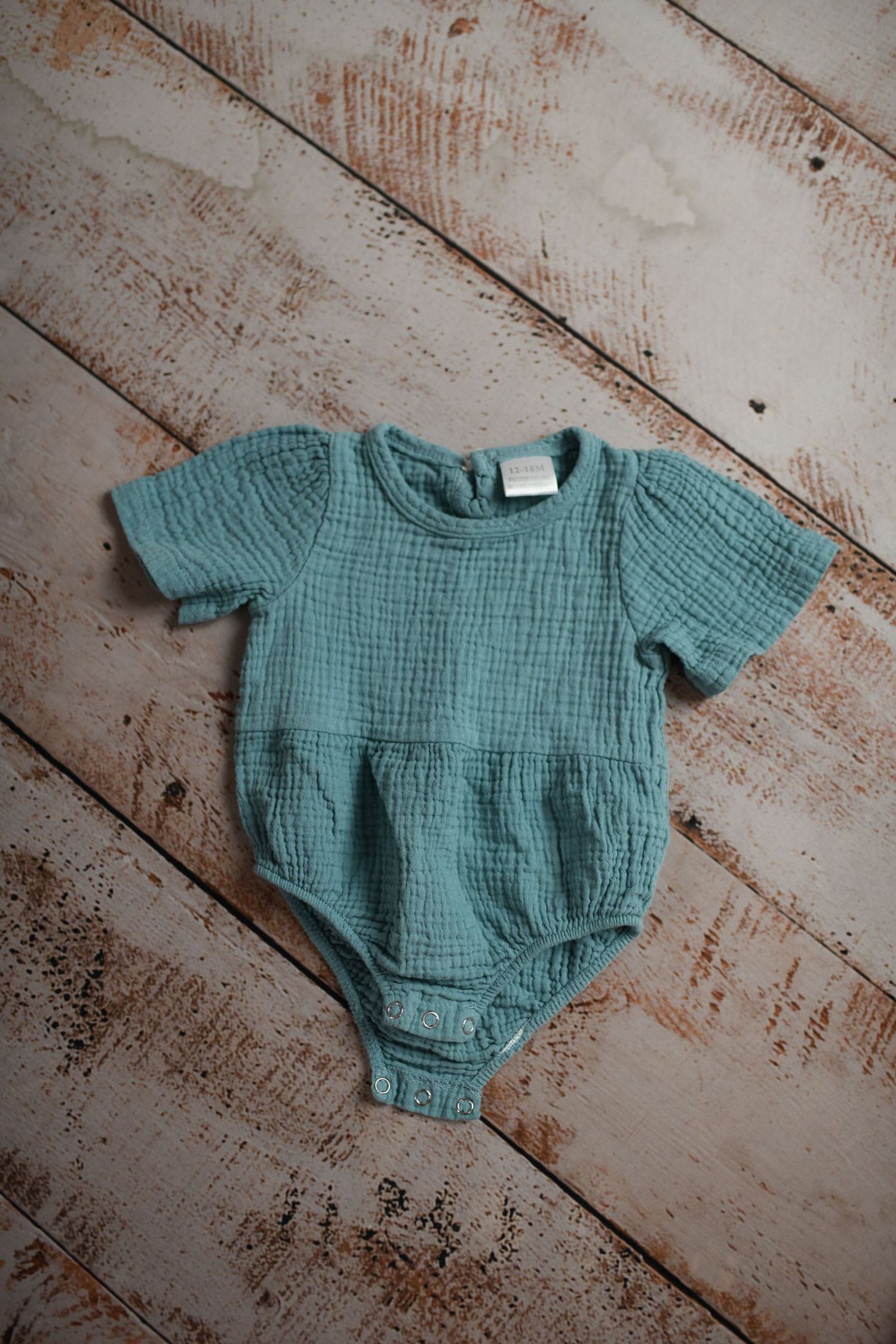 Sage green muslin baby romper displayed on weathered wooden background.