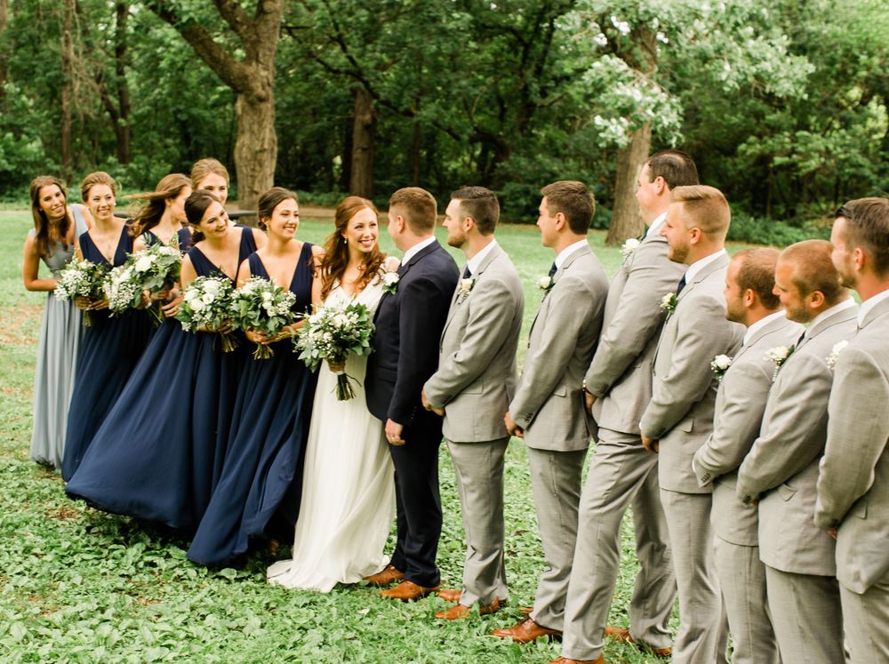 Bride and groom sharing a laugh with wedding party in navy and gray attire under large trees outdoors.