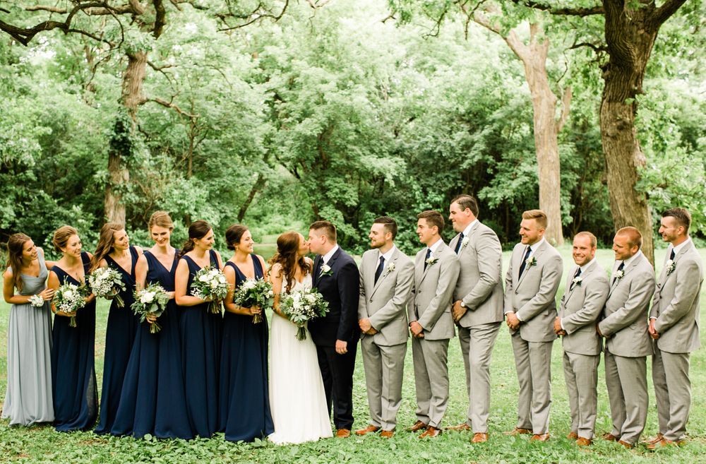 Wedding party sharing a joyful moment outdoors, bridesmaids in navy dresses and groomsmen in gray suits kissing couple.