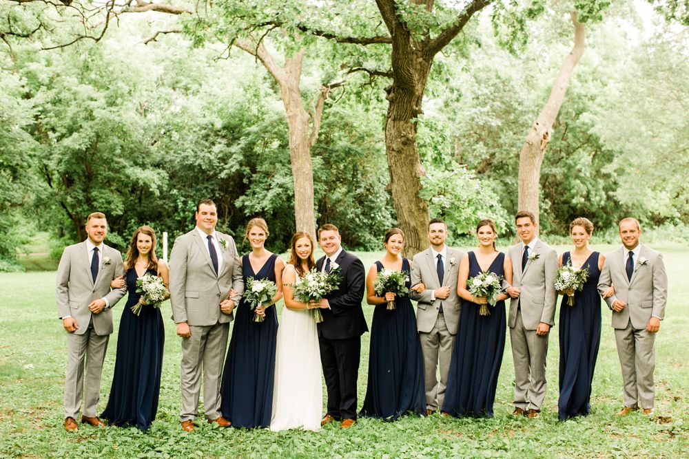 Wedding party smiling together outdoors, bridesmaids in navy blue gowns and groomsmen in tan suits with greenery bouquets.