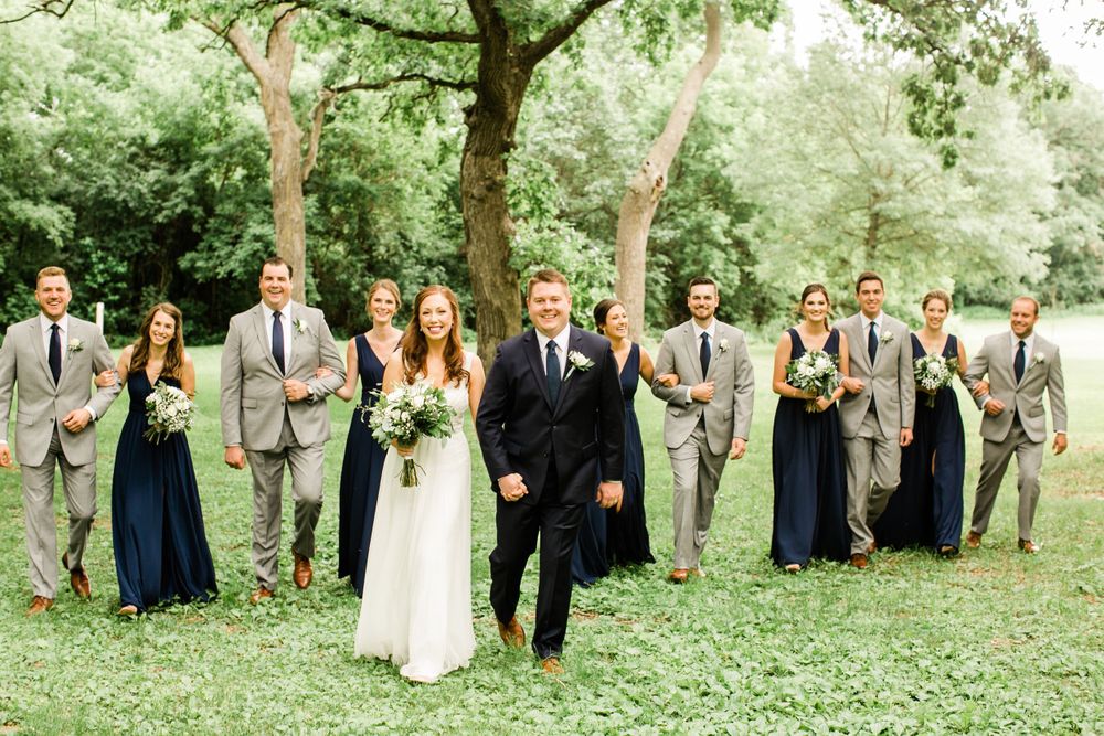Bride and groom walking ahead of wedding party in navy and gray attire through lush green outdoor setting.