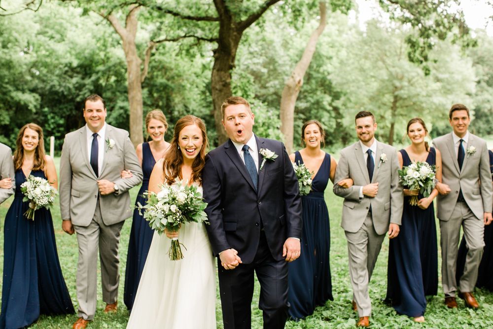 Groom reacting with surprise beside smiling bride, wedding party in navy and gray behind them in green park.