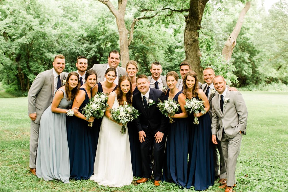 Wedding party group photo outdoors with bridesmaids in navy and gray dresses and groomsmen in gray suits smiling.