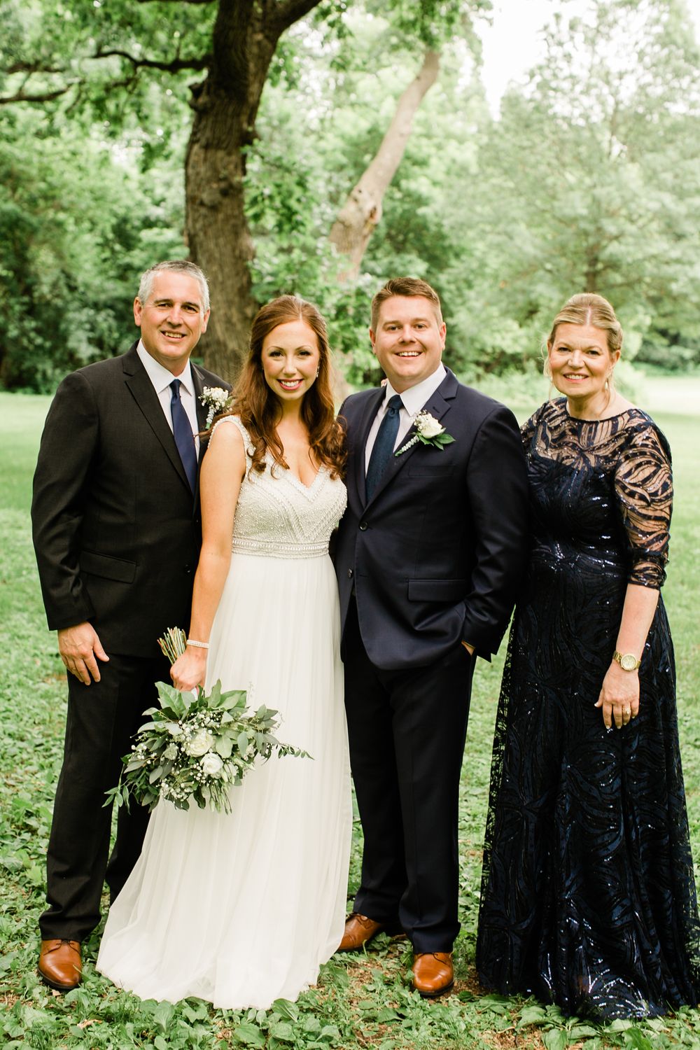 Bride and groom posing with parents outdoors, wearing formal attire including navy suits and lace dress in garden.