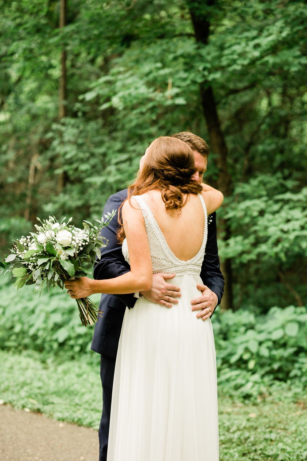 Bride and groom share tender embrace outdoors, bride in elegant backless white gown holding lush greenery bouquet.