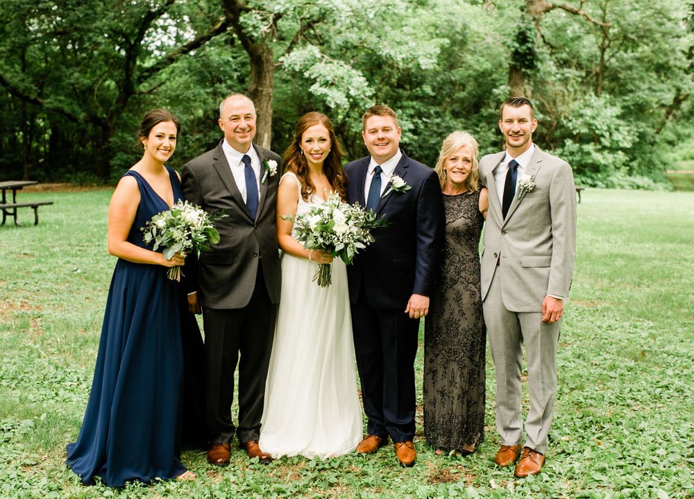 Bride and groom with family members outdoors, wearing navy and gray formal attire with greenery bouquets in garden.