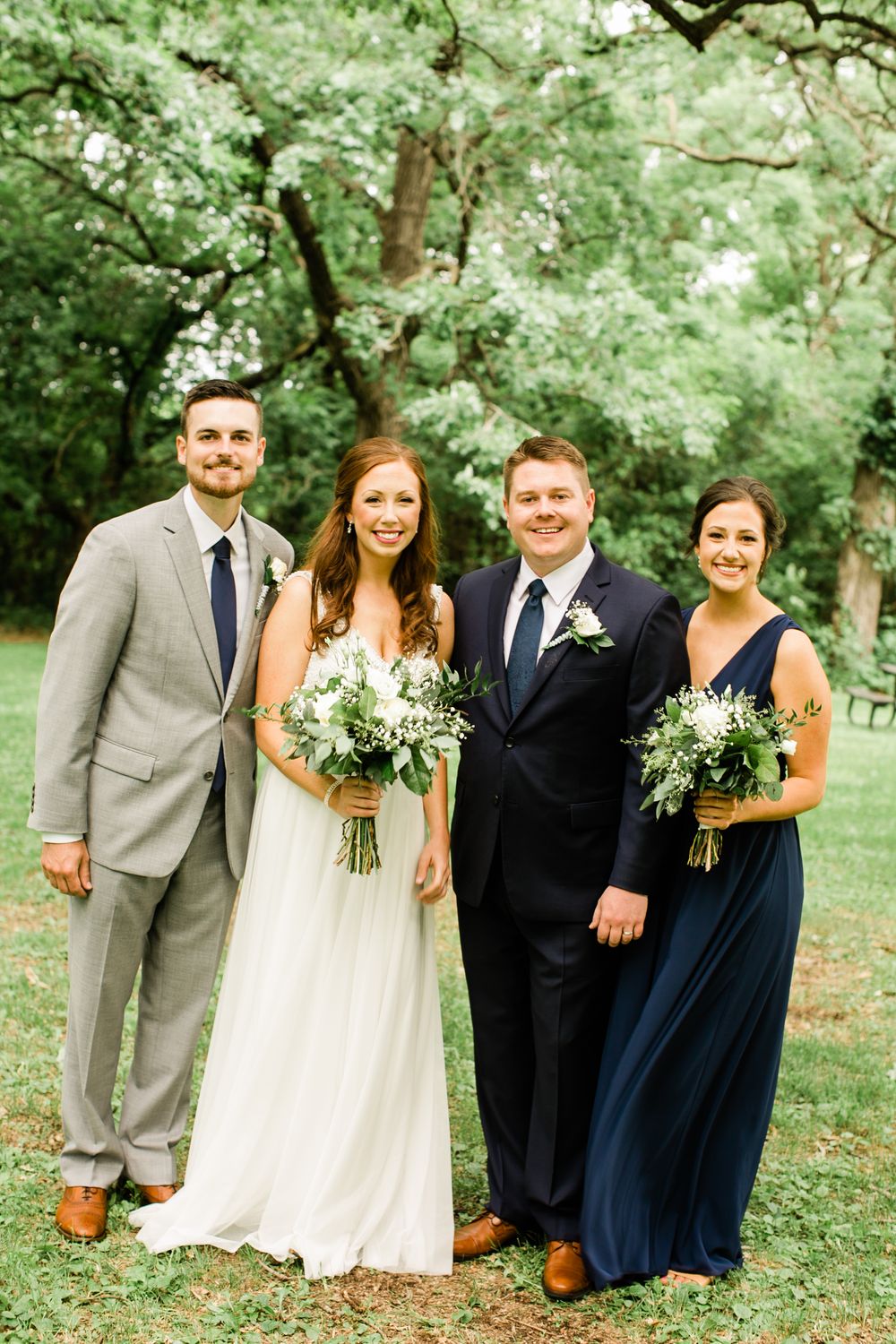 Bride and groom posing with siblings outdoors, wearing navy and gray formal attire with white greenery bouquets.