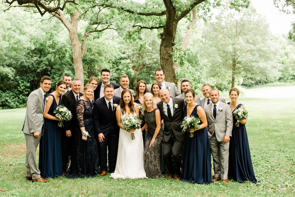 Large wedding party posing together outdoors under a tree, bridesmaids in navy blue dresses and groomsmen in gray suits.