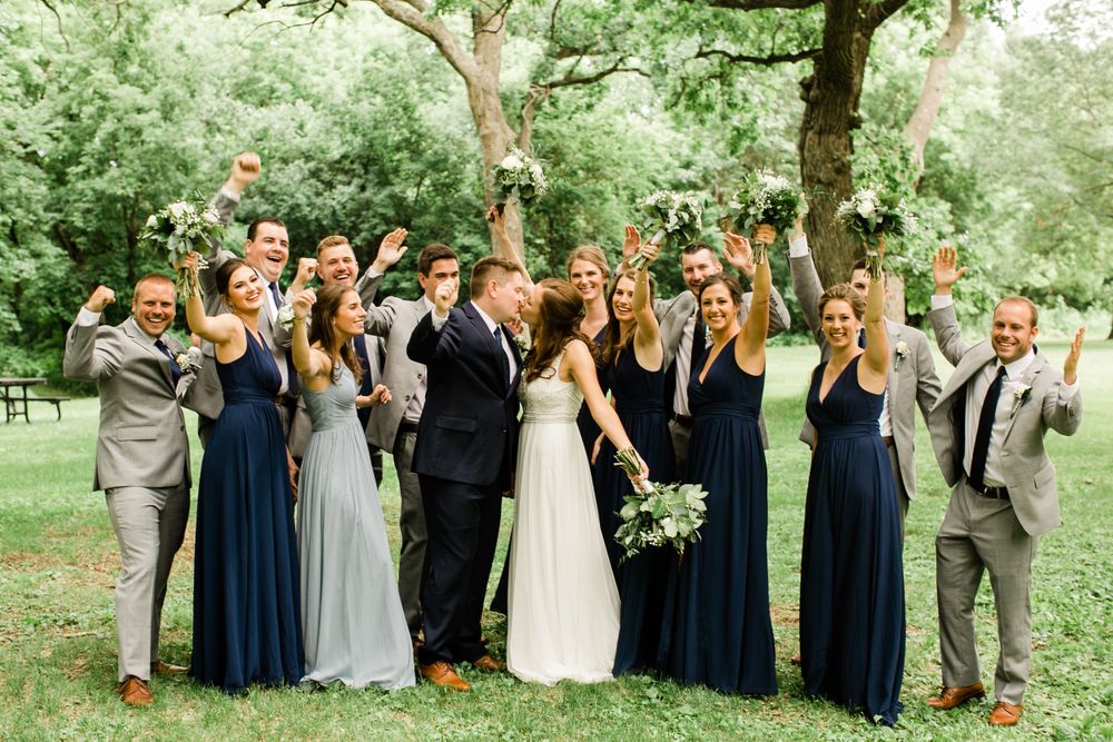 Excited wedding party celebrating outdoors, cheering as bride and groom share a kiss under a large tree.