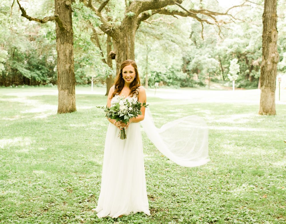 Bride in a flowing white gown holding a greenery bouquet, standing outdoors between two large trees.