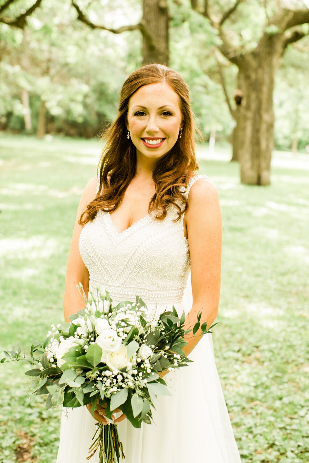 Smiling bride in a beaded white gown holding a lush white and greenery bouquet outdoors under trees.