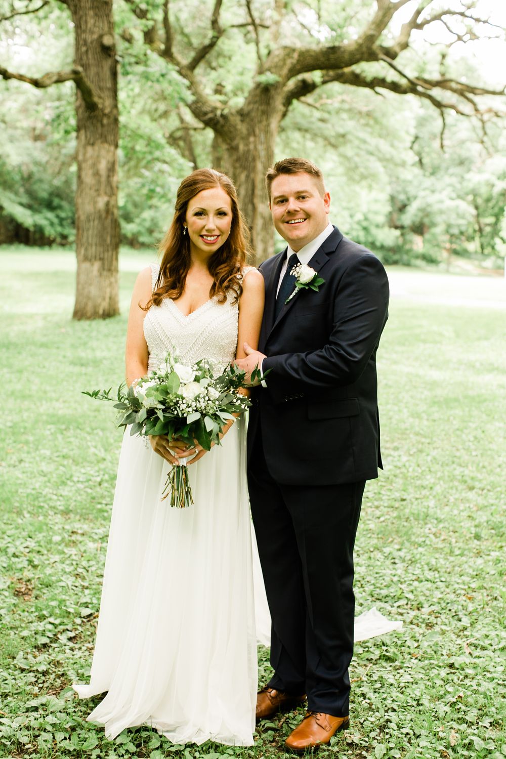 Bride and groom posing together outdoors under large trees, bride holding a white and greenery bouquet.