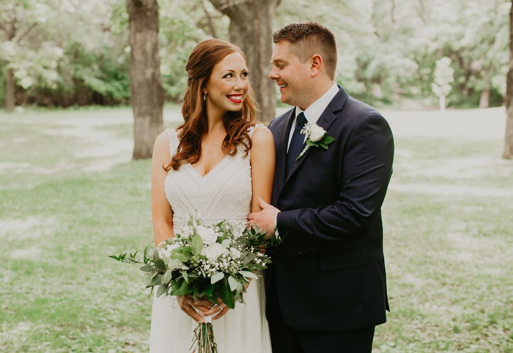 Bride and groom smiling together outdoors, groom in navy suit looking at bride holding a greenery bouquet.