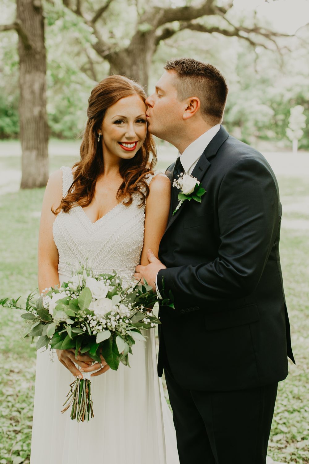 Groom in navy suit kissing bride on the cheek as she smiles, holding a white and greenery bouquet.