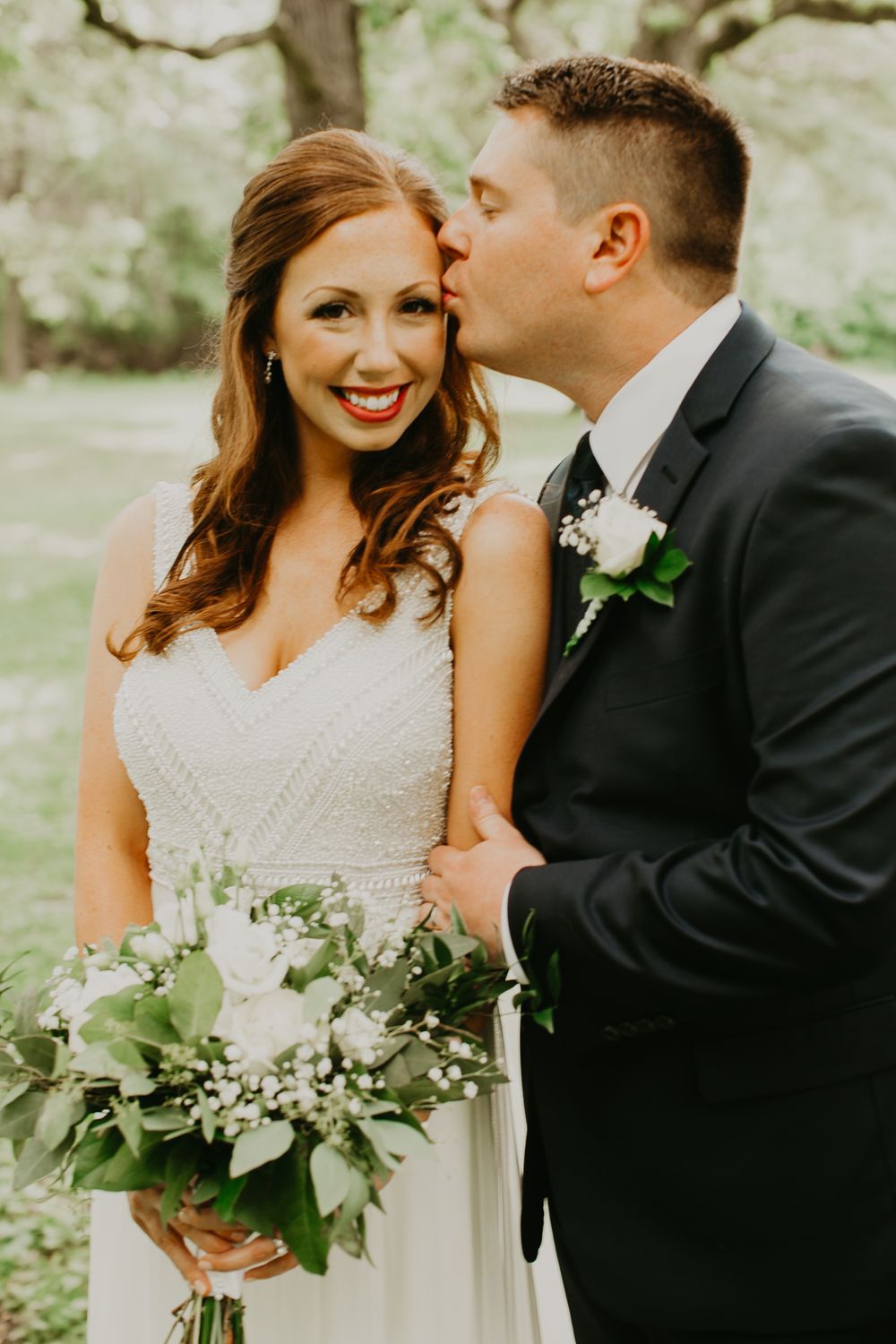 Groom kissing smiling bride on the cheek outdoors, bride holding a lush white and greenery bouquet.