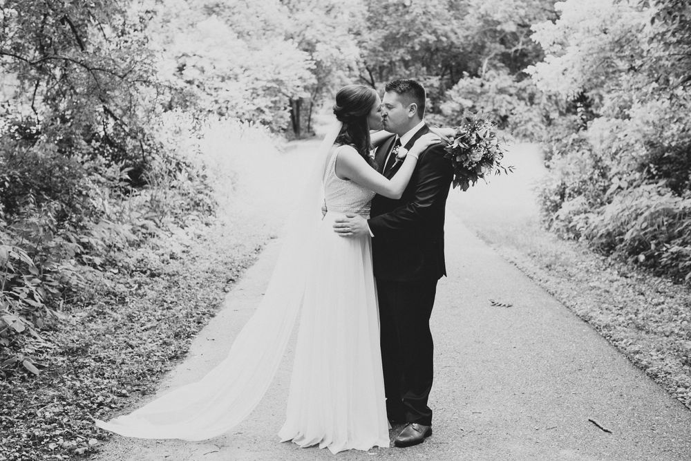 Black and white photo of bride and groom sharing a romantic kiss on a tree-lined pathway.