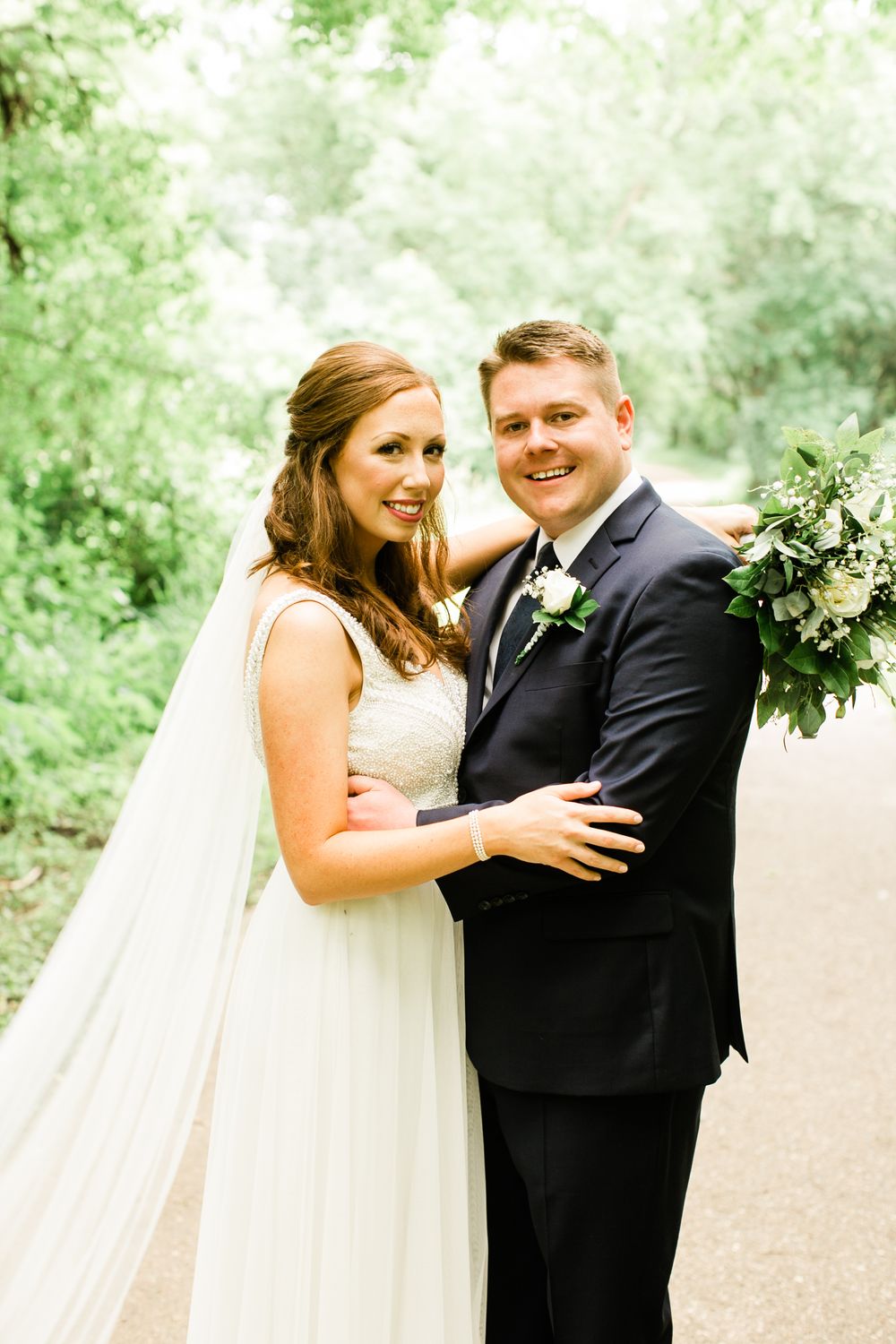 Bride and groom smiling together on a tree-lined path, groom holding a white and greenery bouquet.