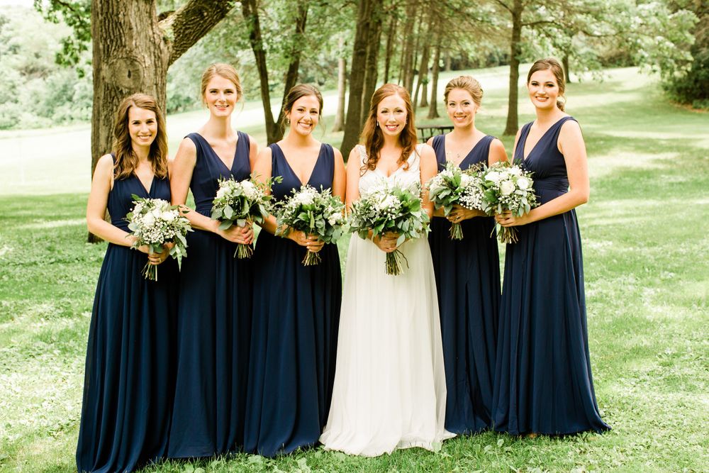 Five bridesmaids in navy blue gowns pose with bride in white dress, holding white floral bouquets outdoors.