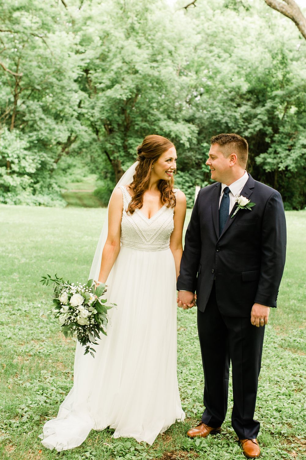 Bride and groom holding hands smiling at each other in a lush green outdoor setting on their wedding day.