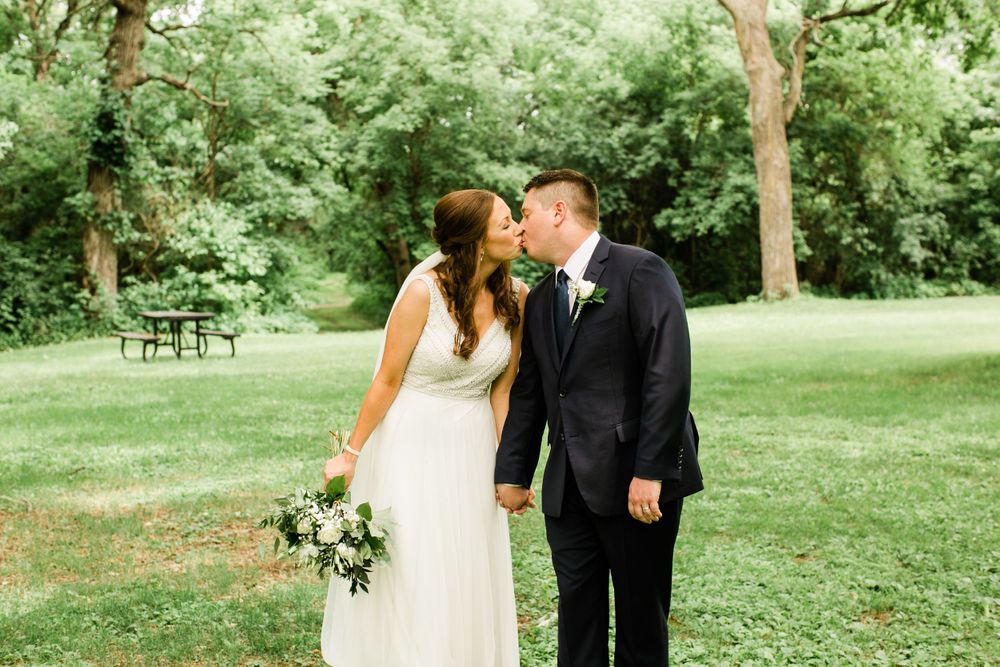 Wedding couple sharing a romantic kiss outdoors surrounded by green trees and grass.
