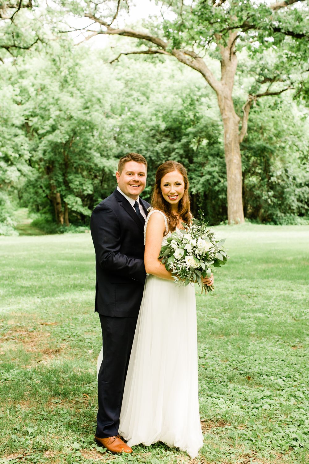 Smiling bride and groom posing together outdoors with lush green trees in the background.