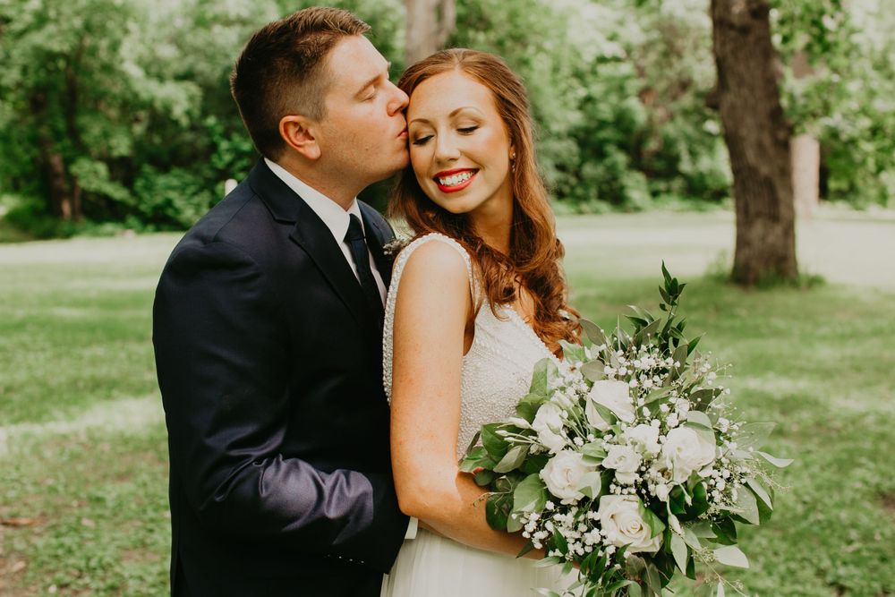 Groom kissing bride on the cheek as she smiles holding a white and green bouquet.