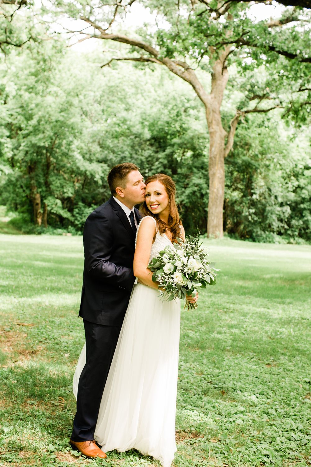 Groom kissing bride on the cheek as she smiles holding a lush bridal bouquet outdoors.