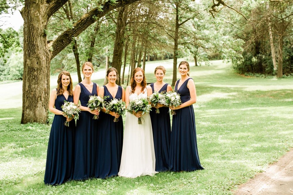 Bride and five bridesmaids in long navy dresses smile together on green lawn surrounded by trees.