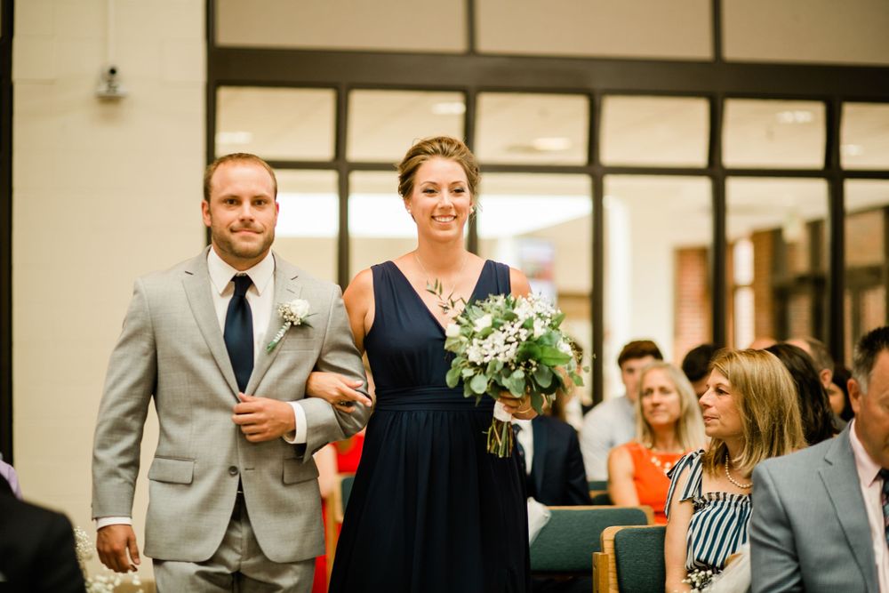 Bridesmaid in navy dress walking down the aisle with a groomsman in a gray suit.