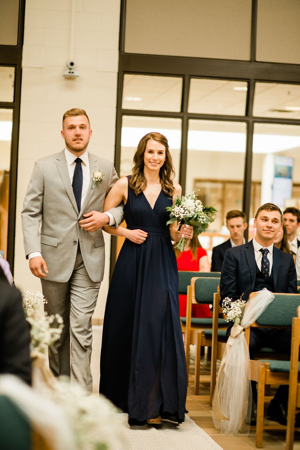 Bridesmaid in navy gown walking down church aisle with groomsman in gray suit at wedding.