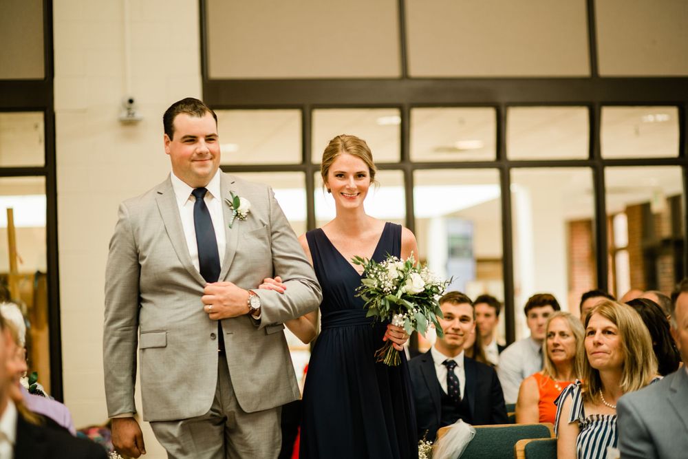 Smiling groomsman in tan suit walks bridesmaid in navy dress carrying white bouquet down ceremony aisle.