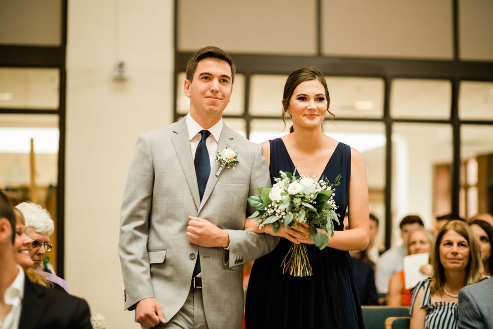 Groomsman in gray suit escorts bridesmaid in navy blue gown down wedding aisle holding white floral bouquet.