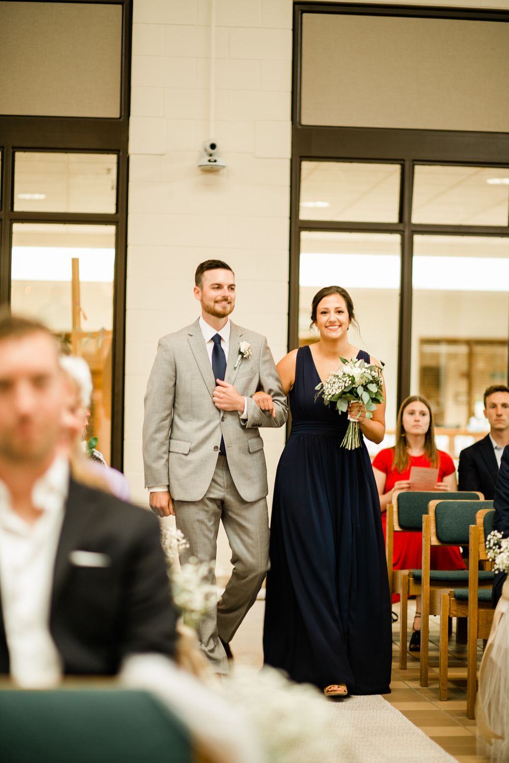 Groomsman in gray suit escorts bridesmaid in navy blue gown down church wedding aisle with guests seated.