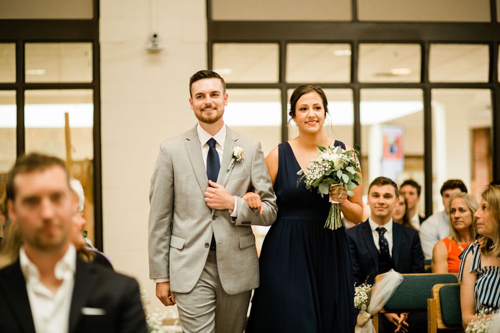Smiling wedding party couple walks down ceremony aisle, groomsman in gray suit with bridesmaid in navy dress.